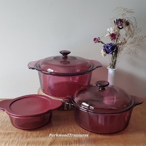 May include: Three vintage Pyrex glass casserole dishes with lids in a deep burgundy color. The dishes are arranged on a wooden surface with a vase of dried flowers in the background.