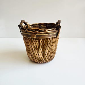 May include: A round, woven brown basket featuring a textured, vertical reed pattern on the body, a reinforced horizontal band near the top rim, and two dark, curved wooden handles attached to the sides of the woven upper border.