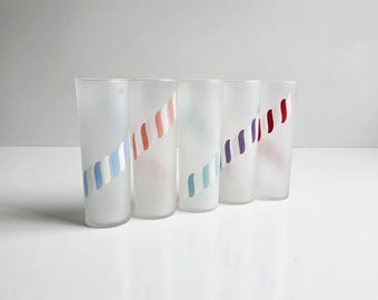 May include: Five frosted glass tumblers with a colorful diagonal stripe design. Each glass features a different color scheme, including blue, orange, teal, purple, and red stripes. The glasses are cylindrical and sit on a white surface.