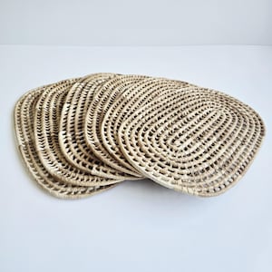 May include: A stack of six oval-shaped woven placemats. The placemats are made of a light tan, natural fiber material. Each mat has an open, lattice-like weave design. The edges are rounded and the mats are stacked on a white surface.
