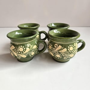 May include: A set of four small olive green ceramic mugs featuring a rounded body and a single handle. Each mug is decorated with a cream-colored floral pattern consisting of swirling vines, leaves, and circular dots around the exterior surface.
