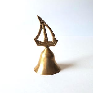 May include: A brass bell featuring a sailboat motif. The bell has a flared base and a decorative sailboat on top. The sailboat design includes lines representing the sails. The bell is a warm gold colour.