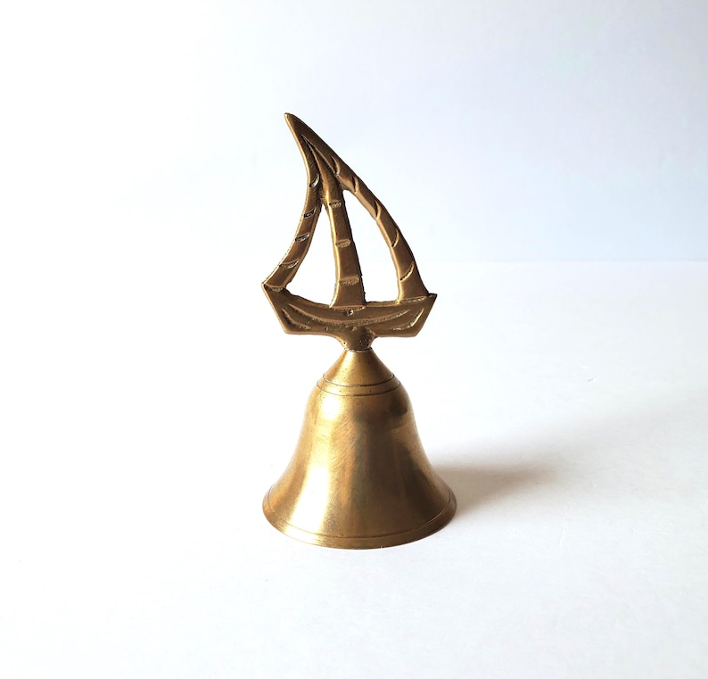 May include: A brass bell featuring a sailboat motif. The bell has a flared base and a decorative sailboat on top. The sailboat design includes lines representing the sails. The bell is a warm gold colour.