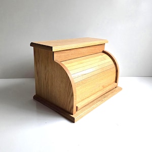 May include: A light brown wooden bread box featuring a roll-top door mechanism. The container has a rectangular base, flat top surface, and curved side panels that frame the slatted sliding door, showcasing a natural wood grain finish throughout.