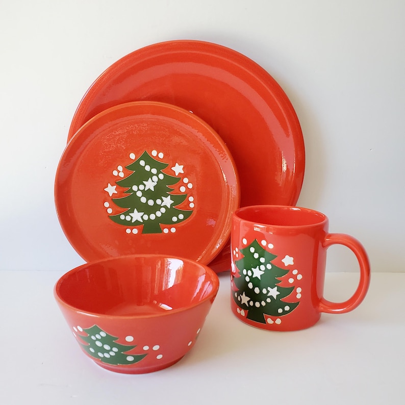 May include: A red ceramic Christmas dinnerware set featuring a large plate, a smaller plate, a bowl, and a mug. Each piece is decorated with a green Christmas tree design and white star accents. The set is perfect for holiday meals.