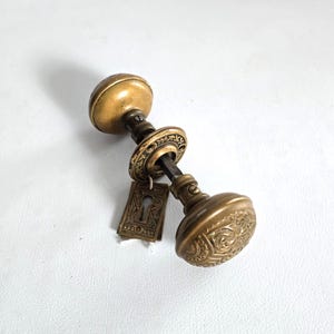 May include: Antique brass door knob set with a keyhole cover. The door knobs have a round shape with detailed floral patterns. The keyhole cover is rectangular with an ornate design. The set is in a vintage style.