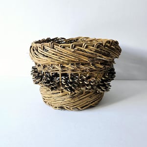 May include: A woven wicker basket with a decorative band of pine cones around the middle. The basket is brown and has a natural, rustic look.