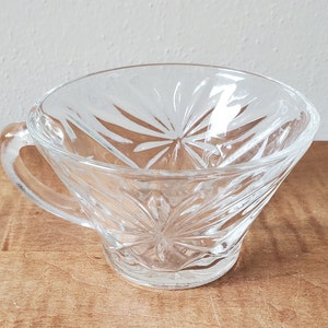 May include: A clear glass cup with a decorative, starburst pattern. The cup has a handle and a wide, shallow bowl.