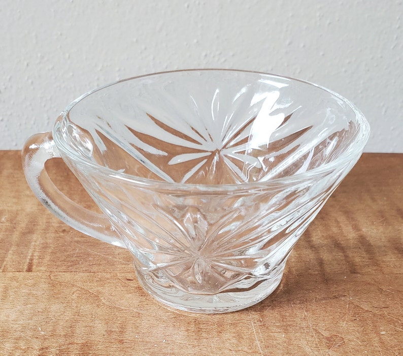 May include: A clear glass cup with a decorative, starburst pattern. The cup has a handle and a wide, shallow bowl.