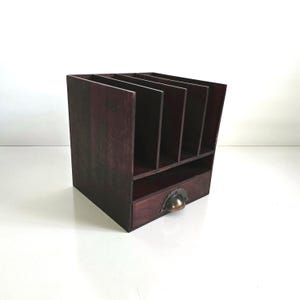 May include: A dark stained wooden desk organizer with multiple vertical slots and a small drawer. The drawer has a decorative metal pull. The organizer is designed to hold papers, files, or office supplies.