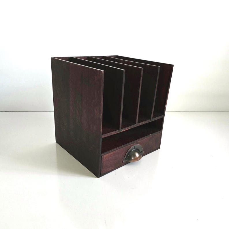 May include: A dark stained wooden desk organizer with multiple vertical slots and a small drawer. The drawer has a decorative metal pull. The organizer is designed to hold papers, files, or office supplies.