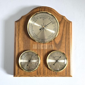 May include: A wooden weather station with three gauges. The top gauge reads "Rain Change Fair" and has a starburst design. Below are a hygrometer and a Fahrenheit gauge, all with gold-colored rims. The wooden backing has a natural wood grain.