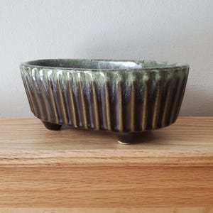 May include: A green and brown glazed ceramic planter with a ribbed design and three small feet. The planter is oval shaped.