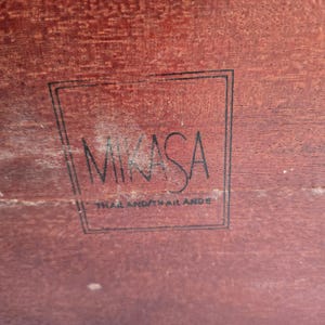 May include: Close-up of a wooden surface with the word "MIKASA" in a black, stylized font, enclosed within a rectangular frame. Below the name, the text "THAILAND/THAILANDE" is printed. The wood has a reddish-brown hue.