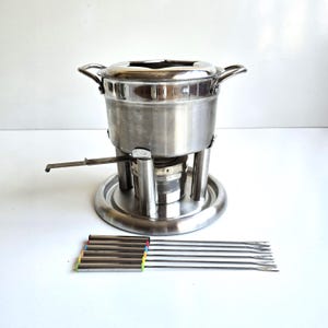 May include: A stainless steel fondue pot with two side handles, a burner stand, and a long adjustment lever. Six metal fondue forks are included, each featuring a distinctively colored end cap in red, blue, orange, yellow, green, and black.