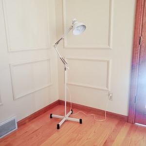 May include: White floor lamp with a flexible arm and a white shade. The lamp is in a corner of a room with wood floors and white walls.