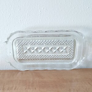 May include: A clear glass butter dish with a decorative diamond pattern and scalloped edges. The dish has two small handles on the sides.