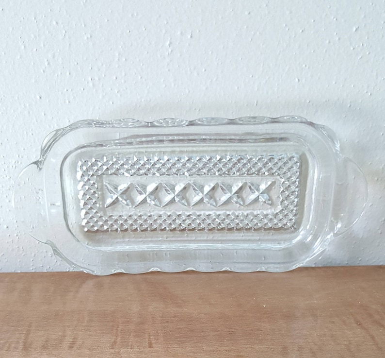 May include: A clear glass butter dish with a decorative diamond pattern and scalloped edges. The dish has two small handles on the sides.