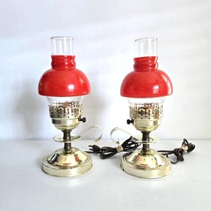 May include: Two vintage table lamps with red shades and clear glass chimneys. The lamps have brass-colored bases and decorative metalwork around the light bulbs. Each lamp has a cord and a switch. The lamps are a classic design.