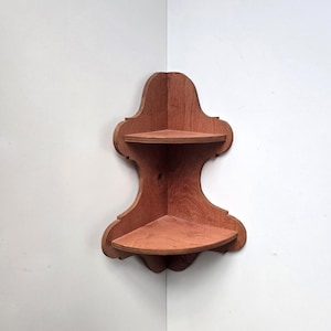 May include: A two-tiered wooden corner shelf featuring a medium brown finish with visible wood grain. The unit has a curved, decorative silhouette with two triangular shelves designed to fit flush against a wall corner for storage.