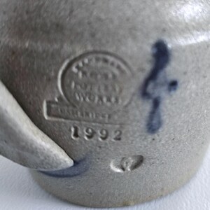 Op de afbeelding: Close-up van een grijs steengoed aardewerk met een handvat. Het stuk heeft een gestempeld merkteken met de tekst "Pottery Works" en "1992". Een blauw decoratief merkteken is zichtbaar aan de zijkant.
