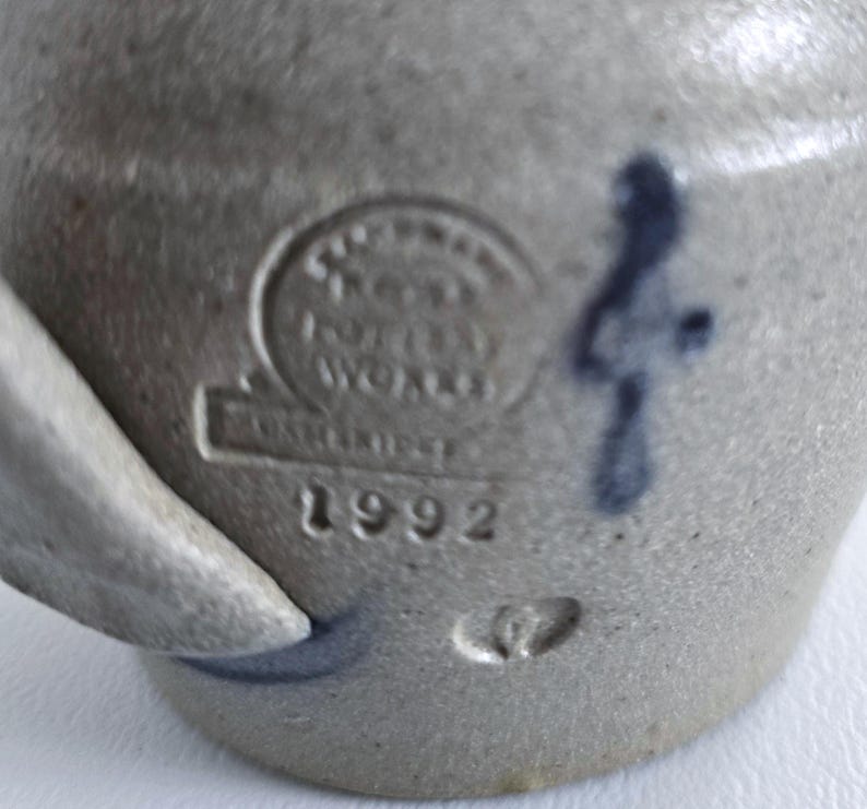 Op de afbeelding: Close-up van een grijs steengoed aardewerk met een handvat. Het stuk heeft een gestempeld merkteken met de tekst "Pottery Works" en "1992". Een blauw decoratief merkteken is zichtbaar aan de zijkant.