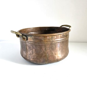 May include: A small, antique copper pot with two brass handles. The pot has a rounded shape and a weathered, aged appearance, with visible patina and discoloration. The pot is likely used for decorative purposes.