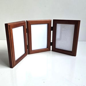 May include: A wooden tri-fold picture frame with a rich brown finish. The frame is hinged, allowing it to stand upright. Each section holds a white photo. The frame is designed to display three photos.