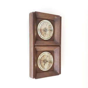 May include: A rectangular wall-mounted weather station featuring a dark brown wooden frame with two circular brass-rimmed gauges. The top gauge displays temperature readings, while the bottom gauge shows barometric pressure with labels including Rain, Change, and Fair.