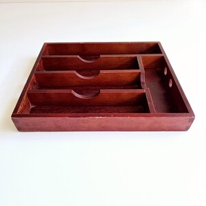 May include: A rectangular, dark-stained wooden desk organiser with multiple compartments. The organiser has three main sections with curved cutouts and a smaller section with two circular holes. The wood grain is visible.