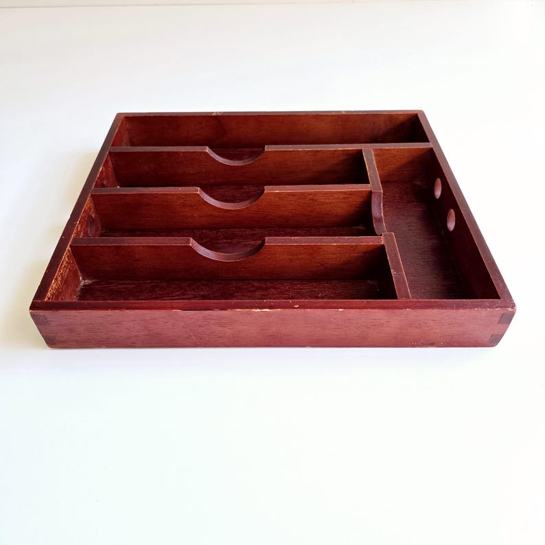 May include: A rectangular, dark-stained wooden desk organiser with multiple compartments. The organiser has three main sections with curved cutouts and a smaller section with two circular holes. The wood grain is visible.