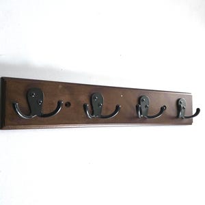 May include: A rectangular dark brown wooden wall rack featuring four black metal hooks. Each hook is secured with two screws and provides two curved arms for hanging items. The wooden base has a smooth finish and a horizontal orientation.