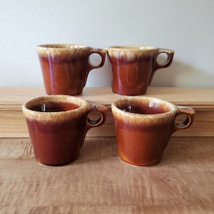 May include: Four vintage brown ceramic mugs with a unique handle design. The mugs have a two-tone color scheme, with a lighter tan rim and a darker brown body. The mugs are arranged on a wooden surface.