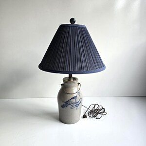 May include: A table lamp featuring a light gray ceramic jug base with a blue hand-painted bird design and a metal wire handle. The lamp is topped with a dark blue pleated fabric lampshade and a small black finial, accompanied by a black power cord.
