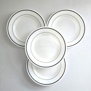 May include: A set of four white ceramic plates arranged in a triangular pattern. Each plate features a smooth, glossy finish and a single, thin, dark green circular band running along the inner rim, creating a clean and uniform aesthetic for dining.