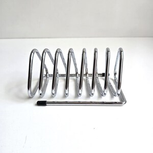 May include: A chrome-plated metal wire rack featuring a series of seven triangular loops arranged in a row. The structure includes a horizontal base frame with small black protective caps on the ends of the wire to prevent scratching surfaces.