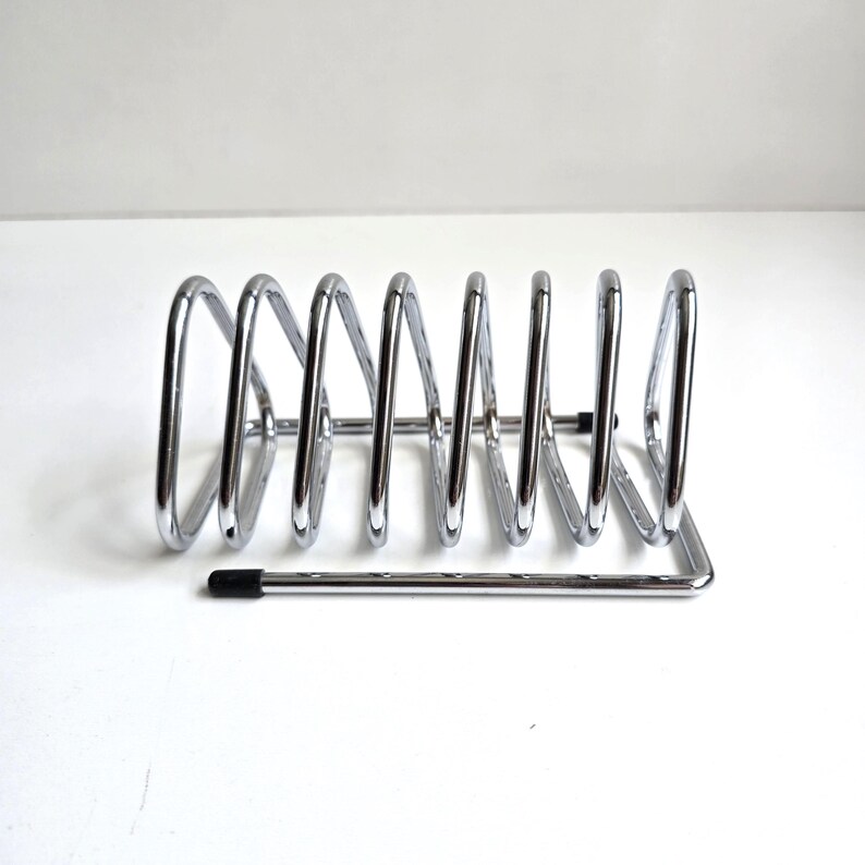 May include: A chrome-plated metal wire rack featuring a series of seven triangular loops arranged in a row. The structure includes a horizontal base frame with small black protective caps on the ends of the wire to prevent scratching surfaces.