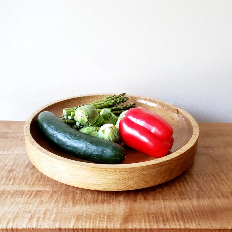 K&ouml;nnte beinhalten: Eine runde, hellfarbene Holzschale gef&uuml;llt mit frischem Gem&uuml;se. Die Schale enth&auml;lt eine gr&uuml;ne Gurke, Rosenkohl, Spargel und eine rote Paprika. Die Schale steht auf einer Holzoberfl&auml;che.