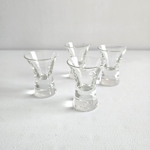 May include: Four clear glass shot glasses with a flared top and a thick, rounded base. The shot glasses are arranged on a white surface, with the light reflecting off the glass.