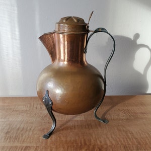 May include: A copper pitcher with a round body, a spout, and a lid. The pitcher has a curved handle and rests on three decorative metal legs. The copper has a warm, reddish-brown hue, and the metal legs and handle are a darker color.