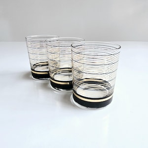 May include: Three clear drinking glasses with black and gold horizontal stripes. The glasses have a cylindrical shape and a thick black band at the base. The stripes create a textured effect, adding a touch of elegance to the glassware.