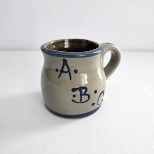 May include: A small, handmade ceramic mug with a rounded body and a handle. The mug is a light beige color with a dark blue rim and handle accent. The letters "A", "B", and "C" are painted in dark blue on the front of the mug.