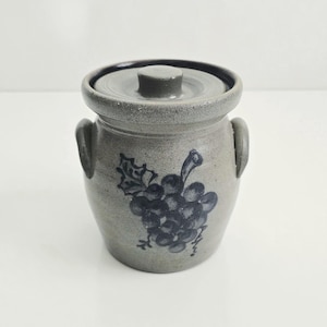 May include: A gray stoneware crock with a lid and two side handles. The exterior features a hand-painted dark blue grape cluster design with leaves. The vessel has a speckled texture and a smooth, rounded rim, providing a functional storage container.