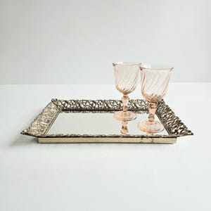 May include: A rectangular mirrored tray with ornate gold-colored filigree edges. Two pink-tinted, stemmed glasses with a spiral design sit on the tray. The glasses reflect in the mirror surface, creating a symmetrical effect.