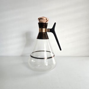 May include: A clear glass carafe with a conical body featuring a metallic copper band around the neck and a matching copper-colored lid. The carafe includes a black plastic handle attached to the neck and a thin copper stripe encircling the lower body.