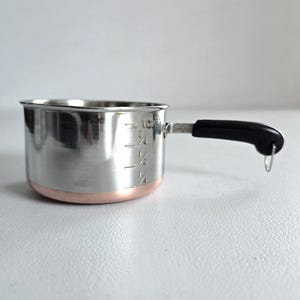 May include: A stainless steel measuring cup with a copper-colored base and a black handle. The cup has measurement markings for 1/4, 1/2, and 3/4 cup, and 1 cup. The handle has a small hole for hanging.