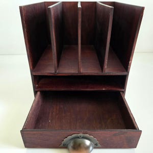 May include: A dark brown wooden desk organizer with multiple compartments and a drawer. The organizer has a vintage aesthetic, with a metal handle on the drawer. The compartments are designed to hold files or other office supplies.