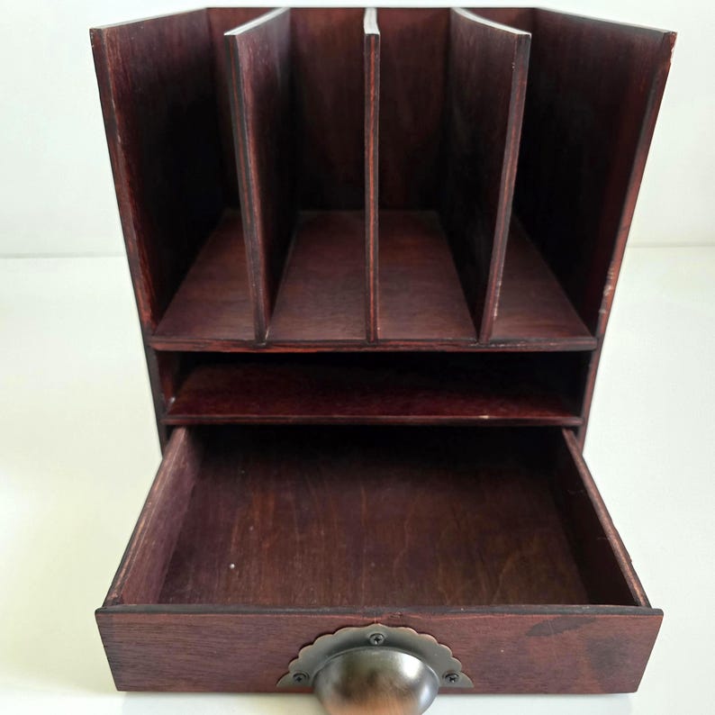 May include: A dark brown wooden desk organizer with multiple compartments and a drawer. The organizer has a vintage aesthetic, with a metal handle on the drawer. The compartments are designed to hold files or other office supplies.