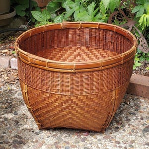 May include: A large, round, woven basket with a natural brown color. The basket has a wide, open top and a sturdy, square base. The weave pattern is intricate and detailed, with a textured appearance. The basket is likely made of bamboo or rattan.
