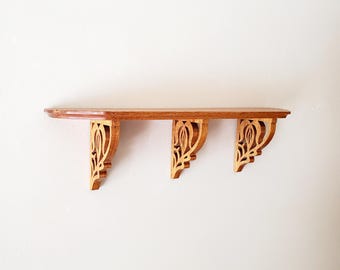 May include: A wooden shelf with three decorative brackets. The shelf is a warm brown color, and the brackets are a lighter shade of wood with a floral design. The shelf is mounted on a white wall.